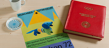 EIn Fotoalbum, eine Tasse und ein Aschenbecher mit Logos der Olympischen Spiele, spwie ein Plakat mit der Aufschrift "München 72" auf einer Tischplatte.