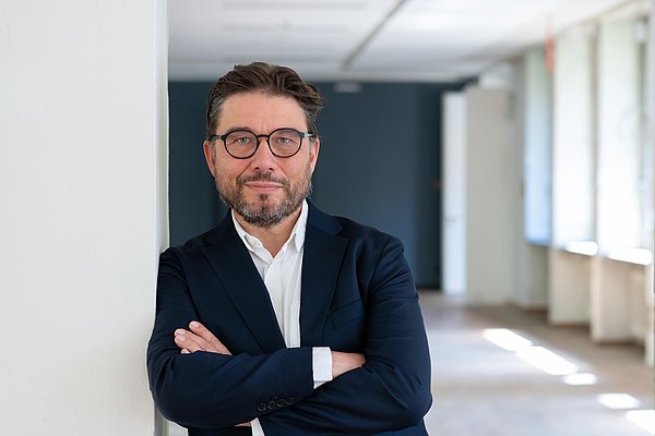 (Dunkelhaariger) Mann mit Brille lehnt an einer Wand mit vor der Brust verschränkten Armen.
