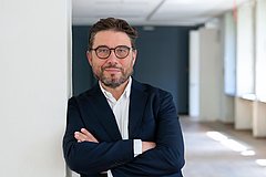 (Dunkelhaariger) Mann mit Brille lehnt an einer Wand mit vor der Brust verschränkten Armen.