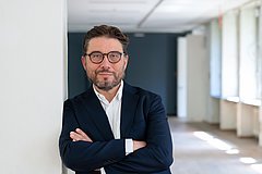 (Dunkelhaariger) Mann mit Brille lehnt an einer Wand mit vor der Brust verschränkten Armen. 