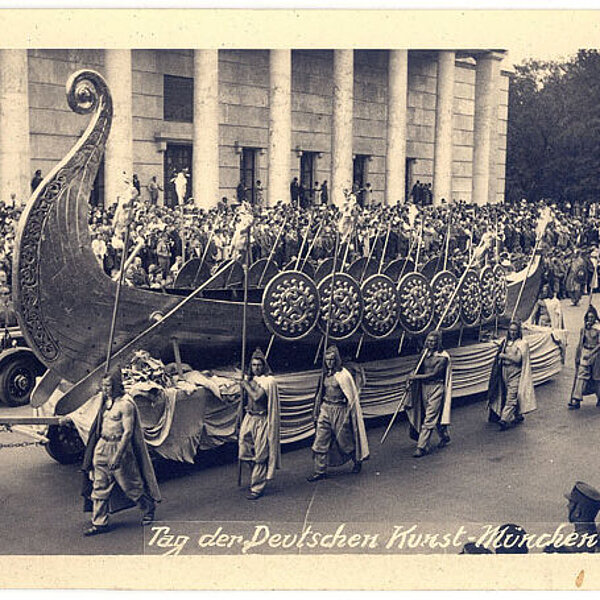 Historische schwarz-weiß-Fotografie eines Festzugs in antik aussehenden Gewändern und nachgebautem, von Pferden gezogenen hölzernen Kriegsschiff.