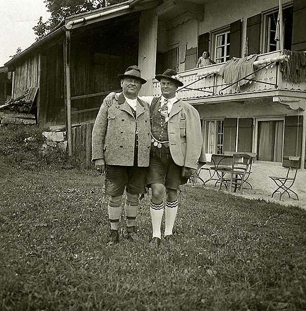 Historische Schwarz-weiß-Fotografie zweier Männer in bayerischer Tracht vor einem Bauernhaus.