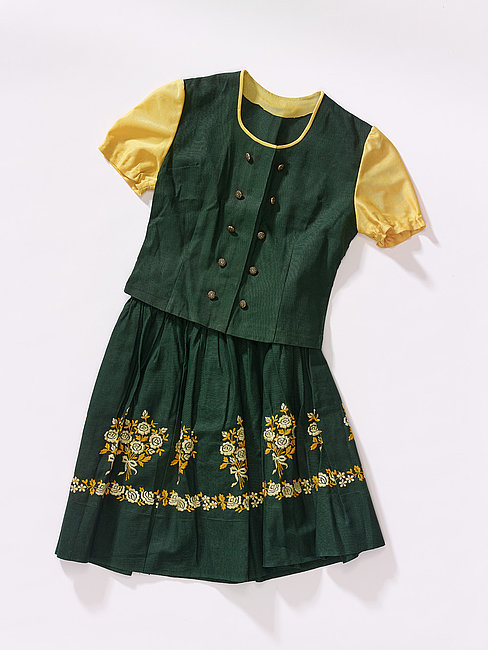 Das Foto zeigt ein Kinderdirndl aus dunkelgrünem Stoff mit aufgestickten gelben Rosen auf dem Rock und gelben Puffärmeln.