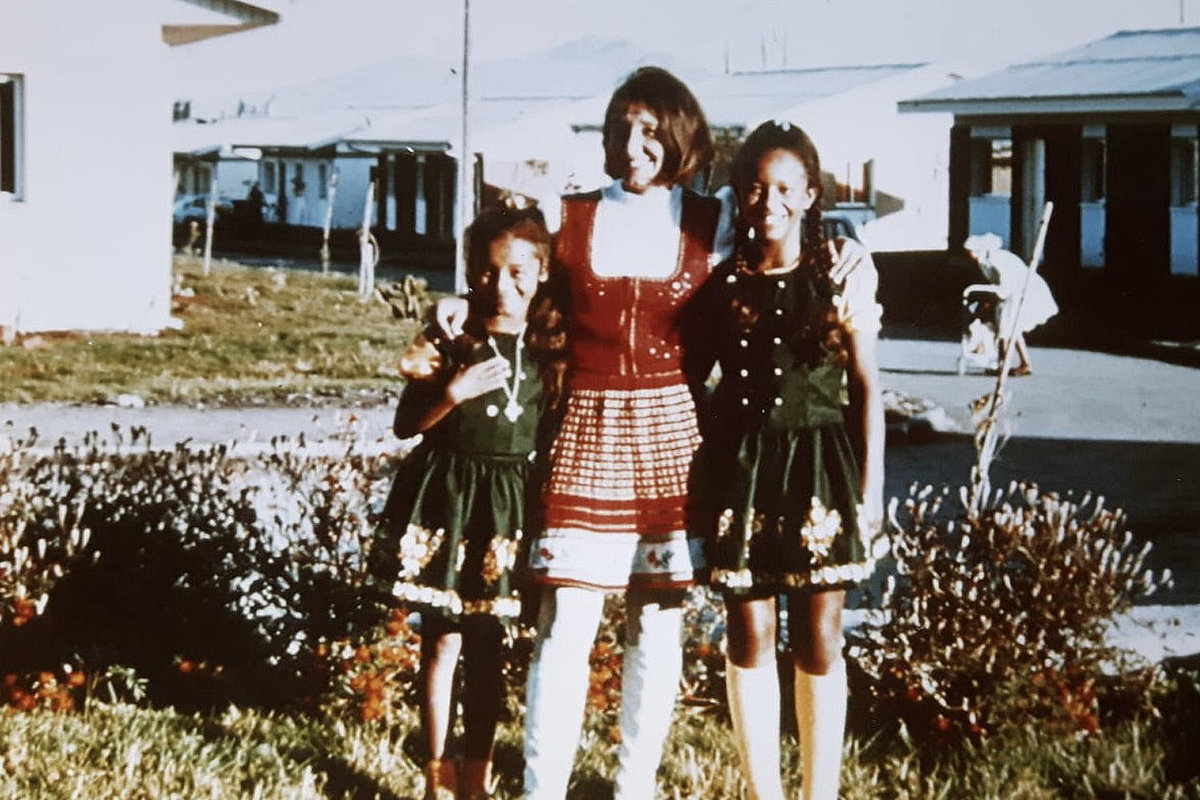 Eine lachende Frau in rotem Dirndl hält ihre beiden jungen Töchter in grünen Dirndlkleidern rechts und links im Arm.