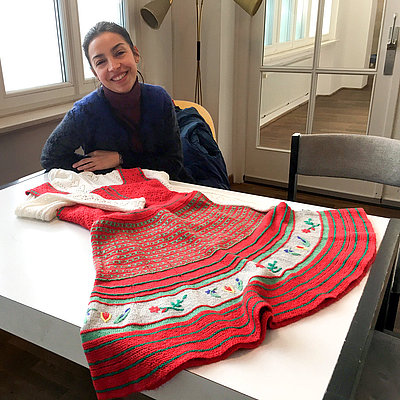 Eine lächelnde junge Frau sitzt an einem Tisch, auf dem ein Dirndl liegt, gestrickt aus roter, grüner und weißer Wolle.