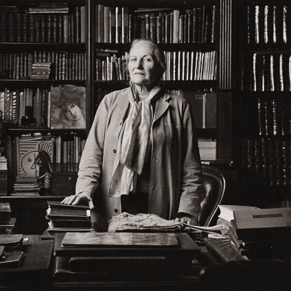 Frau von 80 Jahren in grauem Arbeitskittel am Schreibtisch stehend, im Hintergrund ein hohes Bücherregal