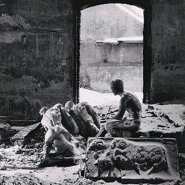 Historische schwarzweiß-Fotografie mehrerer beschädigter Statuen in einem zerstörtem Raum voller Trümmer.